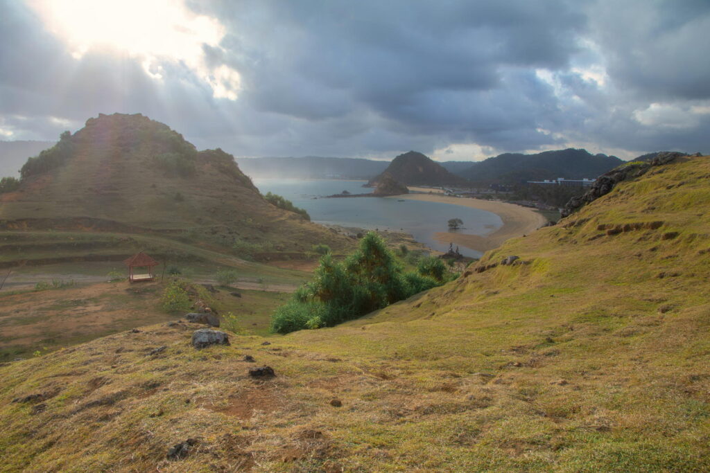 A coastal landscape featuring a sandy bay surrounded by lush green hills under a cloudy sky, with sunlight breaking through – one of the many stunning things to do in Lombok.