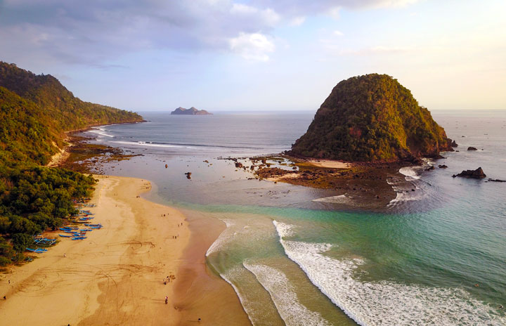 Aerial view of an empty sandy beach with clear waters, small waves, and green vegetation on the left. Two hills rise from the ocean, one prominently near the shore and another farther out—one of many picturesque spots among things to do in Banyuwangi.