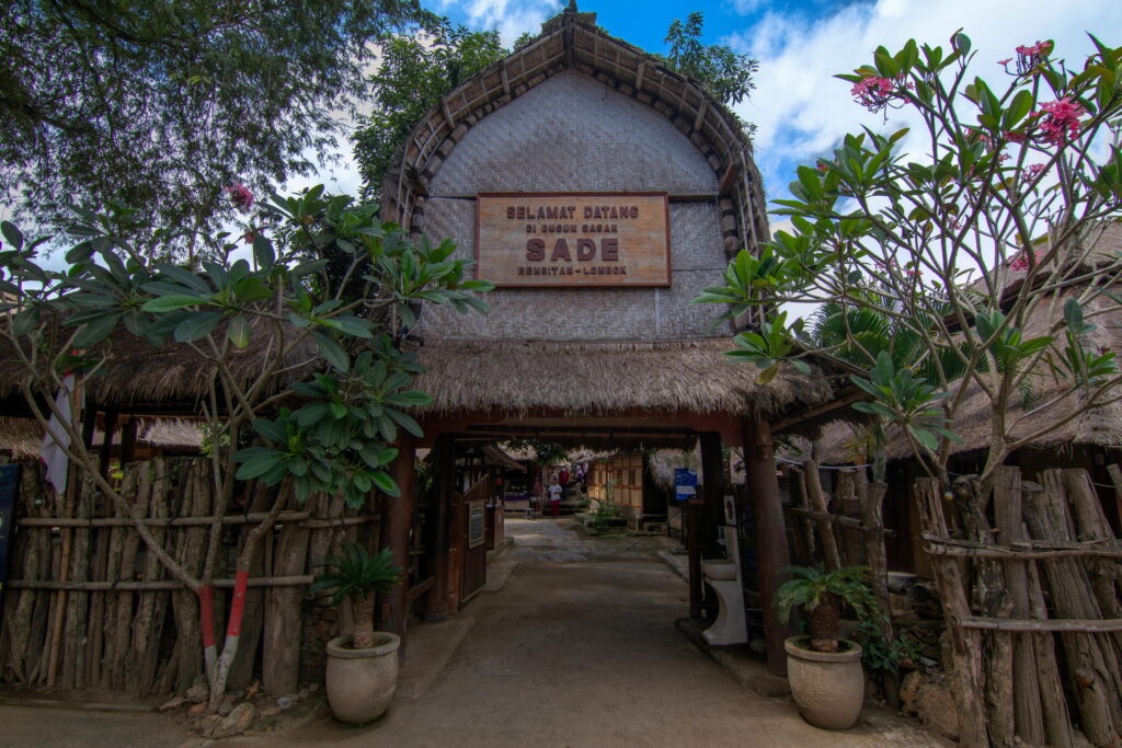 Entrance to a traditional village with a thatched roof gate, wooden fence, and green foliage. The sign above the gate reads “Selamat Datang di Desa Sade,” welcoming visitors to Sade Village. Among the things to do in Lombok, exploring Sade offers a glimpse into local culture and traditions.