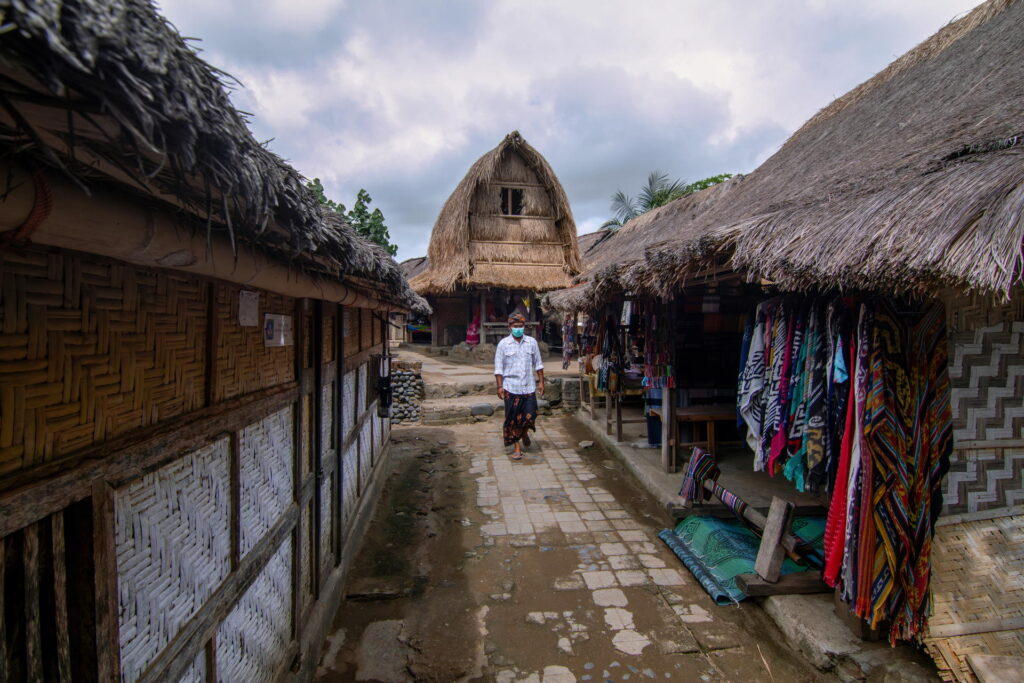 A person walks down an alley with traditional thatched-roof buildings and market stalls under a cloudy sky, discovering the charm and local culture of things to do in Lombok.