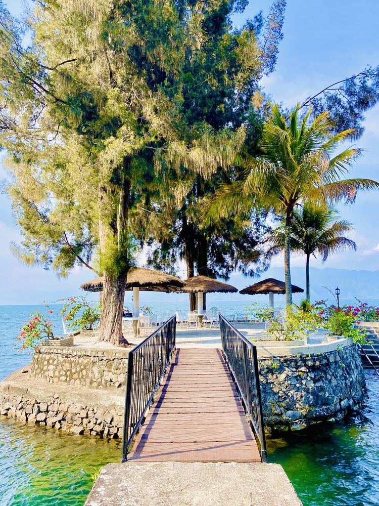 A wooden walkway leads to a small, tree-covered island with wicker umbrellas and stone walls, nestled in the tranquil expanse of Lake Toba.