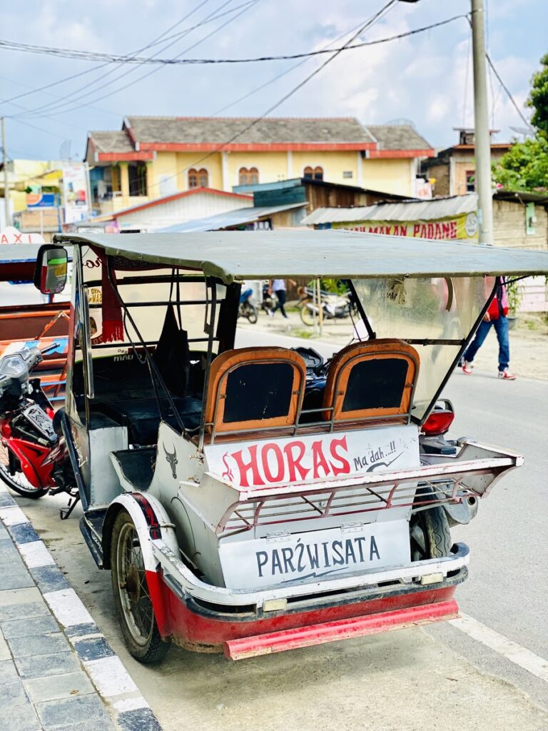 A motorized tricycle with a "HORAS" sign and the word "PARIWISATA" on the back is parked on a street, capturing the lively spirit of Lake Toba, surrounded by buildings and power lines.