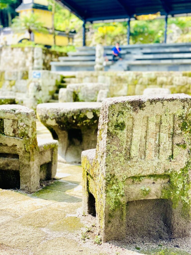 Stone benches and steps in an outdoor seating area near the tranquil shores of Lake Toba, where moss-covered surfaces merge seamlessly with lush greenery in the background.