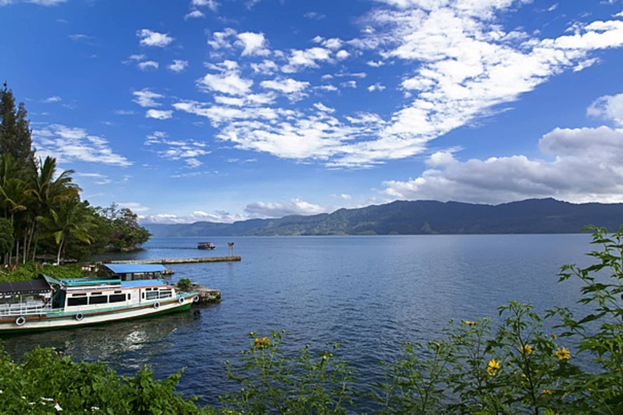 Lake Toba, with its serene waters and a small docked boat, is surrounded by lush green vegetation and majestic mountains under a partly cloudy sky.