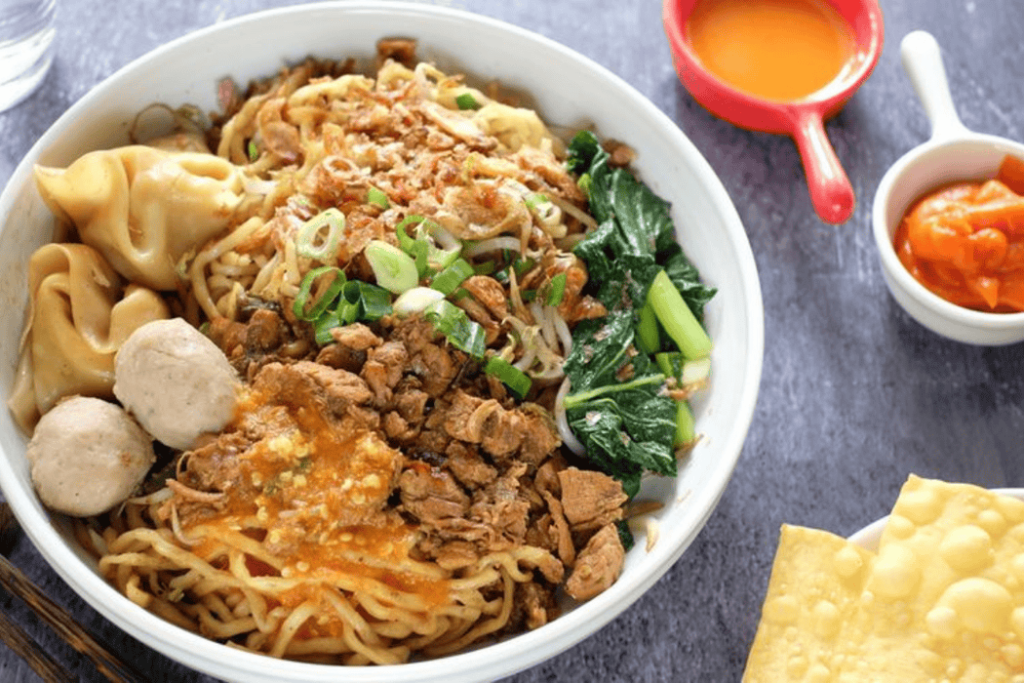 A Yogyakarta-inspired noodle soup with dumplings, meatballs, greens, chopped meat, scallions, sauces, and crispy wontons for a delightful experience.