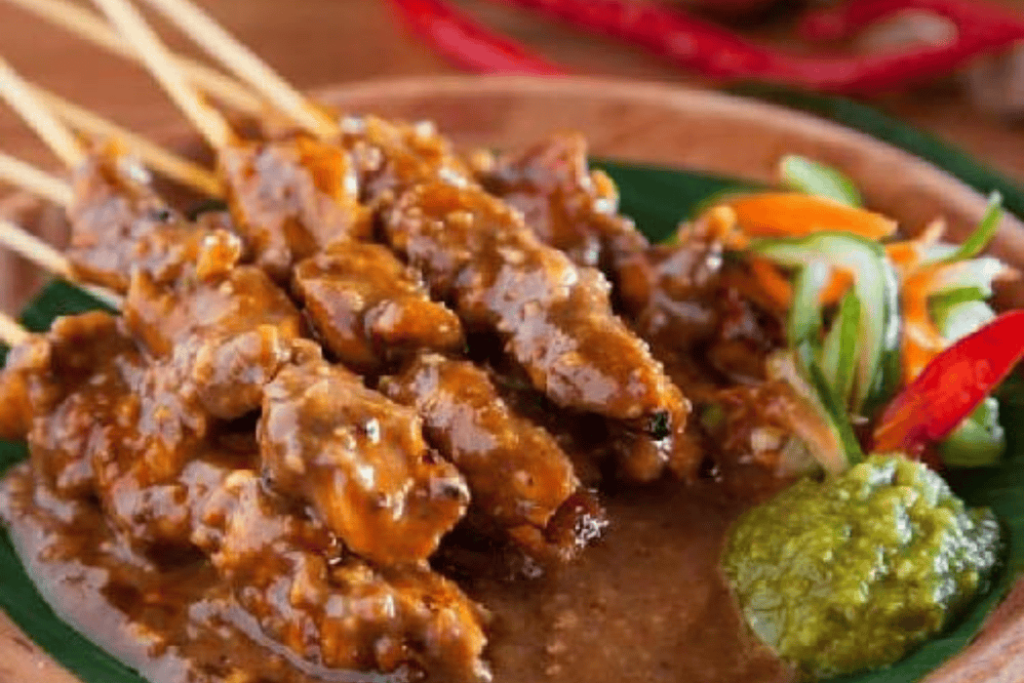 A plate of Yogyakarta-style meat skewers with peanut sauce, garnished with sliced vegetables and a dollop of green sauce.