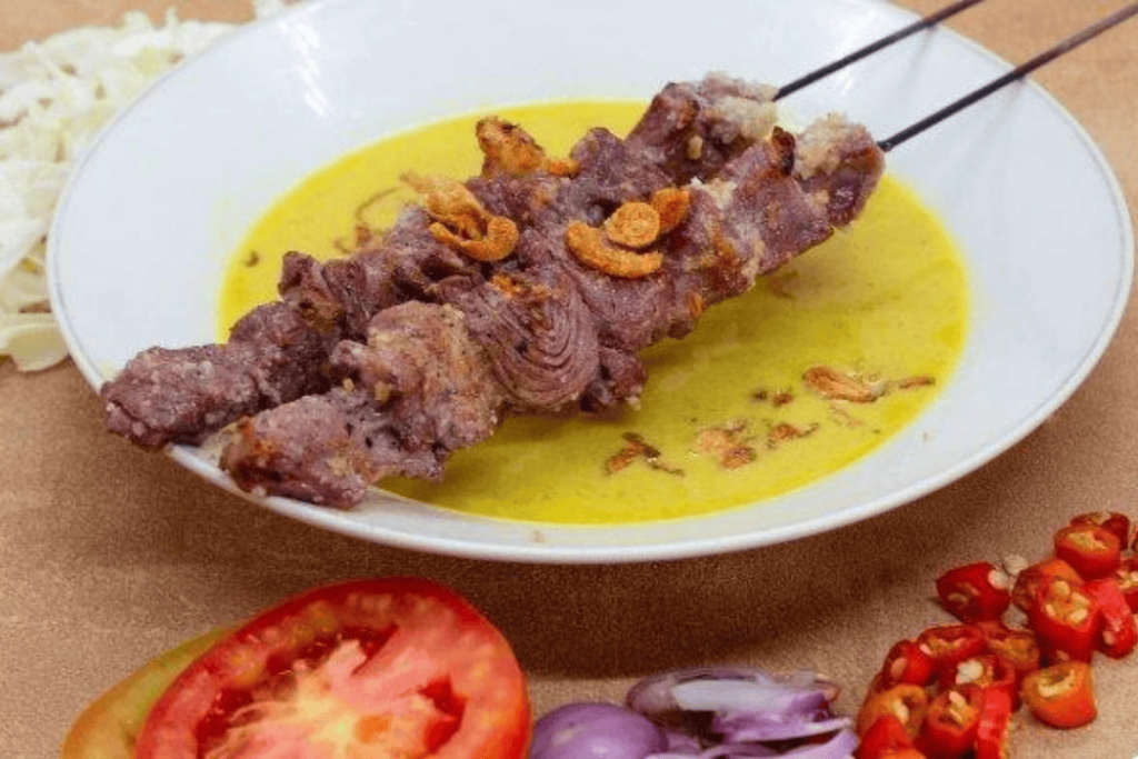 Two grilled meat skewers in yellow sauce, garnished with garlic, chili, tomato, and shallots, showcase Yogyakarta's vibrant flavors.