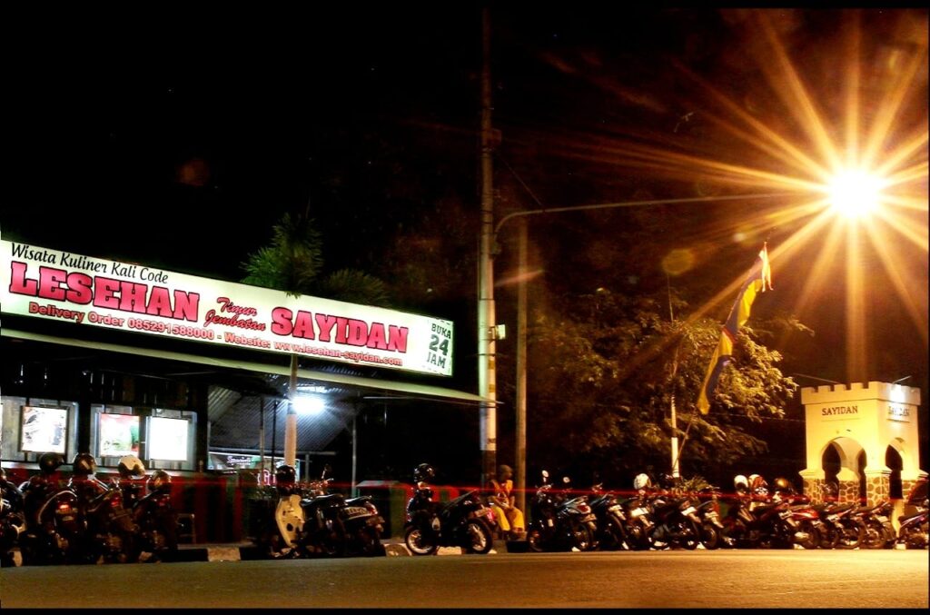 Under a Yogyakarta streetlight, a vibrant food stall shines, surrounded by parked motorcycles and the bustling energy of the lively night scene.