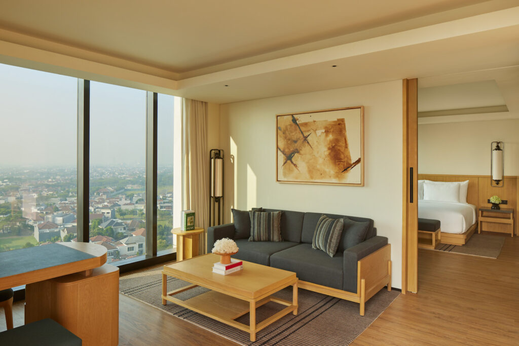Modern Hotel Tentrem Jakarta suite with a living area featuring a gray sofa, wooden furniture, and city view through large windows; a bedroom is partially visible through sliding doors.